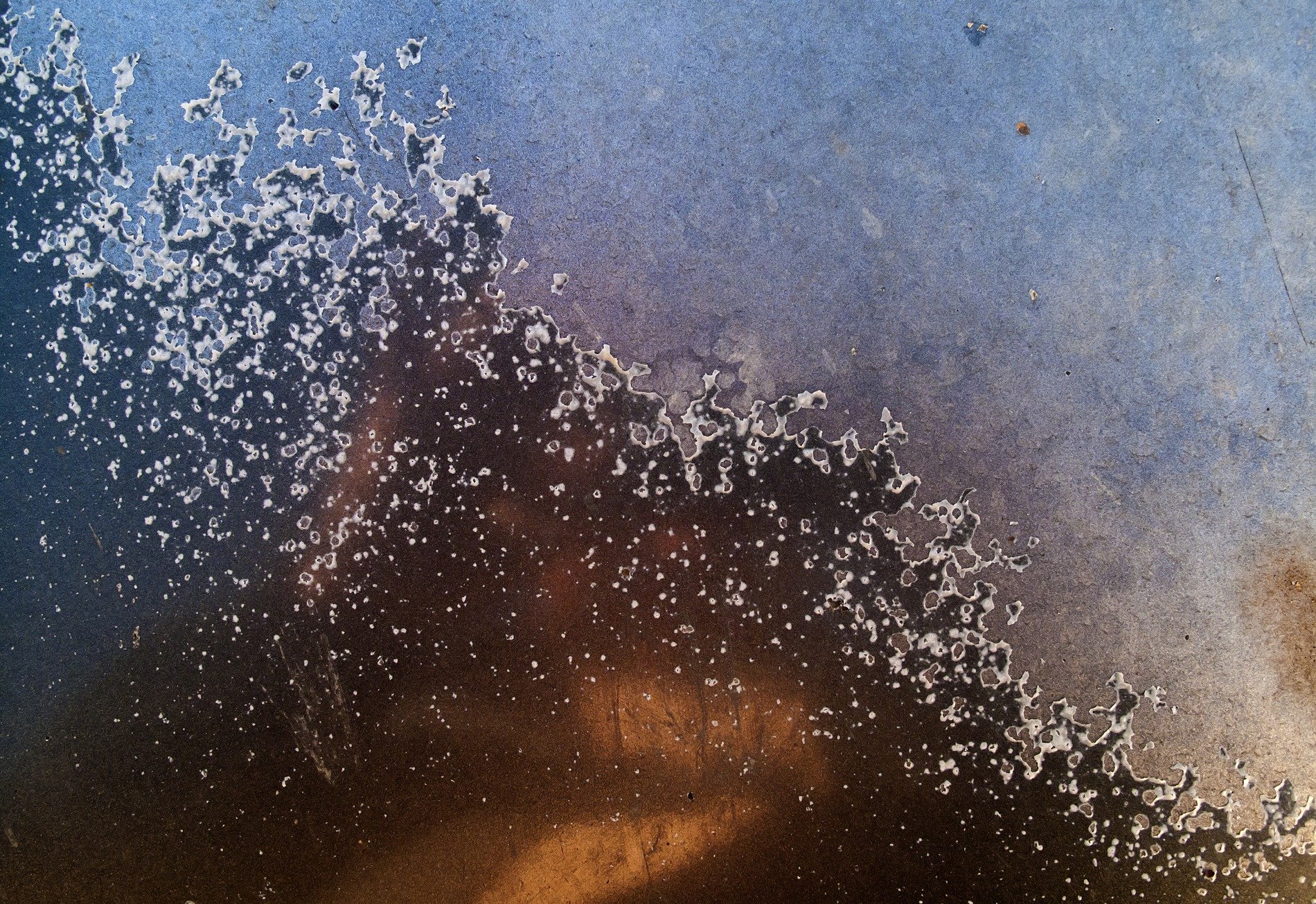 Blue fields meet the dark, white specks line the rusted edge, grit marks the shoreline