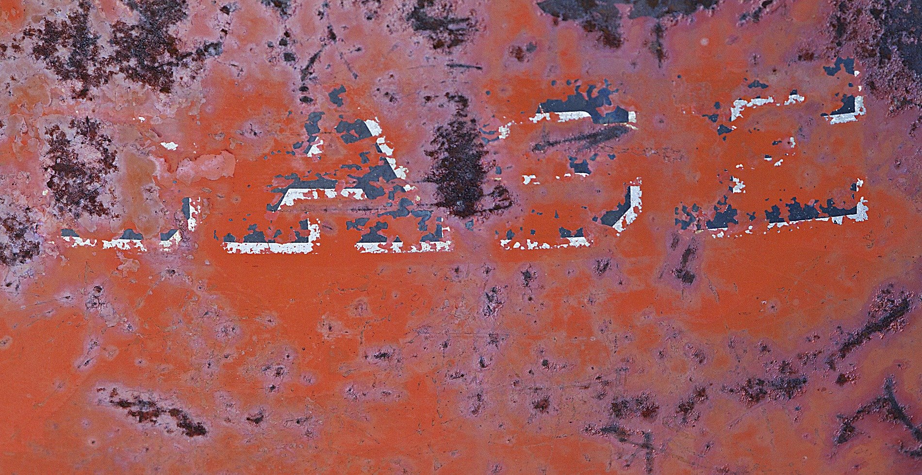 Red rust eats the sign, white paint peels from pitted steel, letters fade away