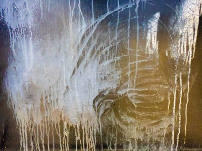 Blue flakes drift away, light catches the jagged edge, grit marks the clear pane