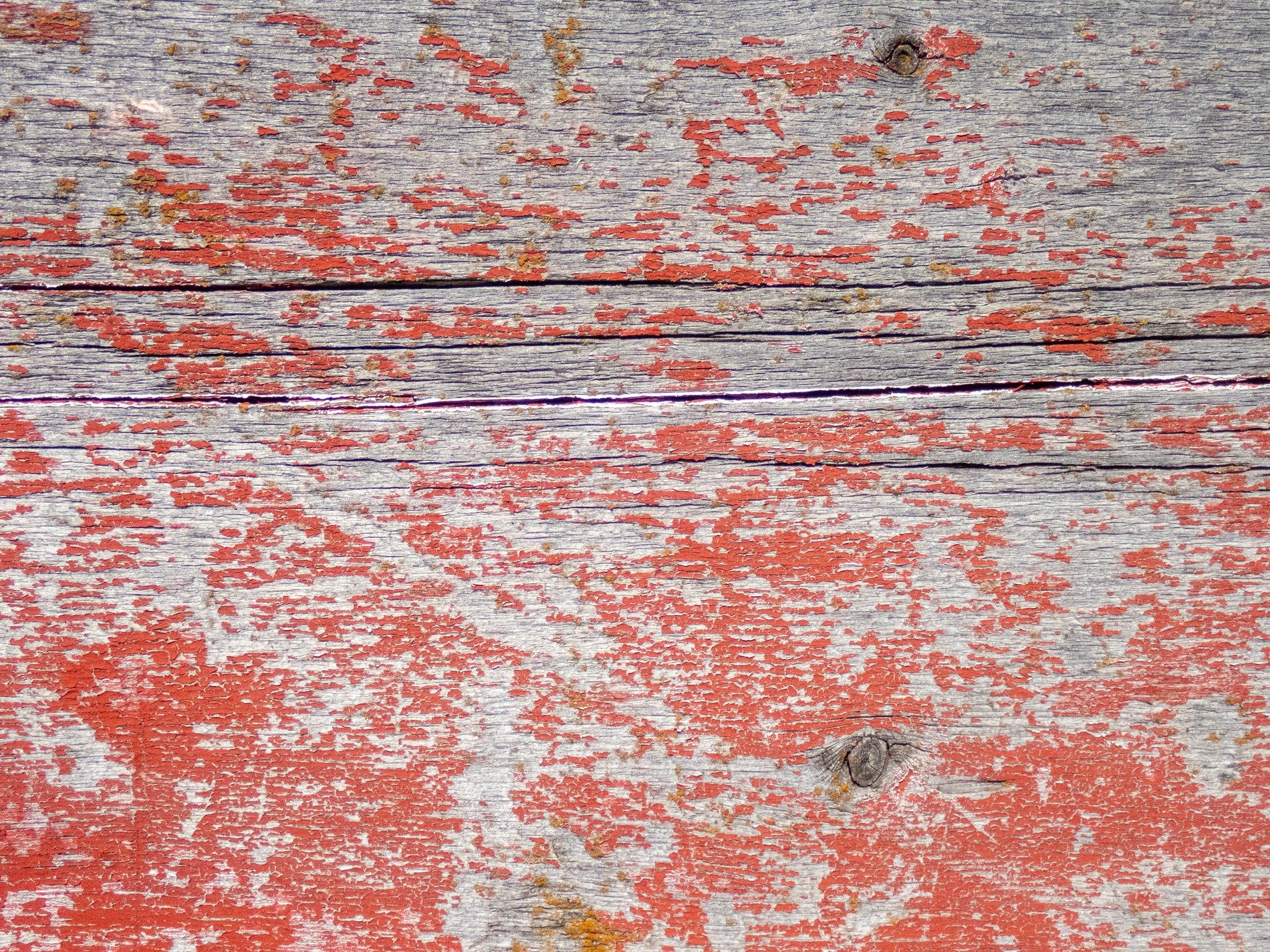 Red paint flakes away, grey wood grain reveals its age, cracks split through the plank