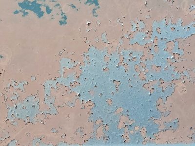 Blue paint curls away, tan metal hides underneath, time peels back the layers