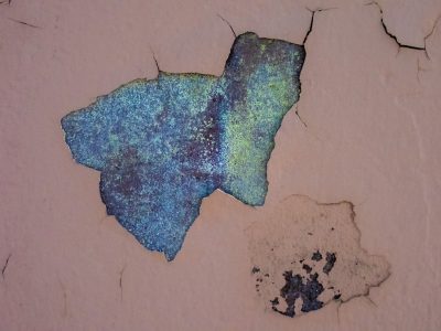 Pale paint curls away, azure depths bloom underneath, spider cracks reach out