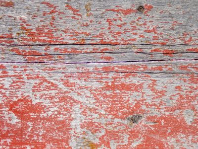 Red paint flakes away, grey wood grain reveals its age, cracks split through the plank
