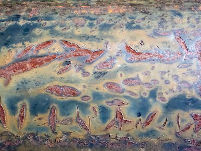 Teal paint peels away, red rust blooms in jagged gaps, weathered metal skin