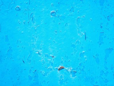 Bright blue paint curls back, rusted metal waits beneath, flakes drift like thin ice